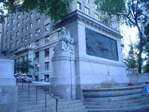 A large stone and metal memorial with a brick apartment building behind it.