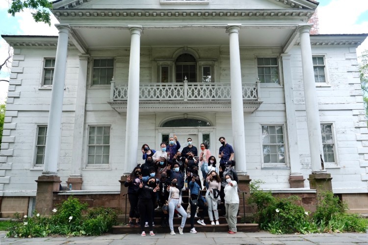 Photo by Henry Danner. A group of high school students posing on the steps in front of a large white house.