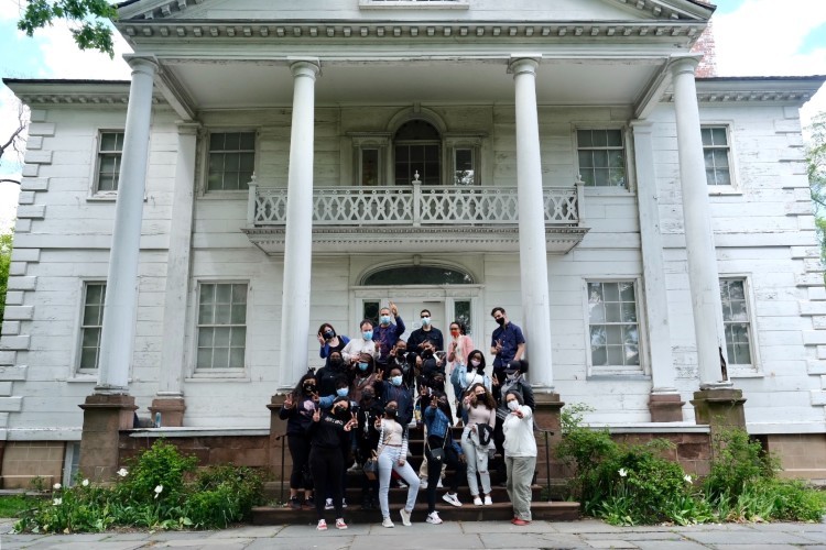 A group of high school students posing on the steps in front of a large white house.