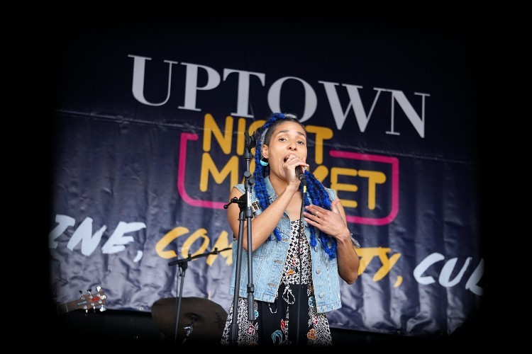 A woman singing into a microphone in front of an Uptown Night Market banner