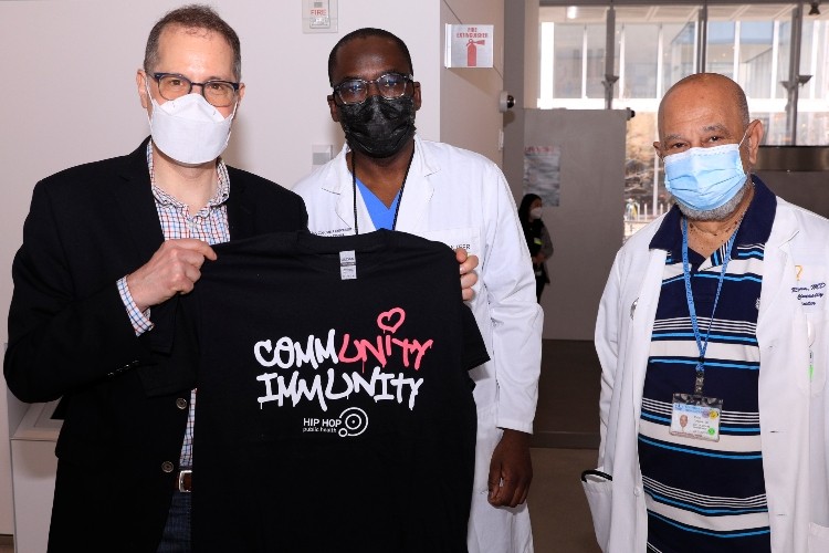 New York City Council member Mark Levine, Dr. Olajide Williams, and Dr. Rafael Lantigua in the lobby of The Forum. Levine holds up a black Community Immunity t-shirt that highlights the word Unity 