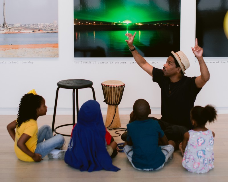 A man speaks to a group of children about art that's visible behind him.