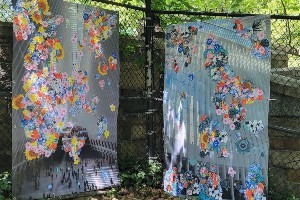 A picture of two works of art leaning against a chain link fence. The artworks are photographs of the Oculus in Lower Manhattan with flowers stitched on them.