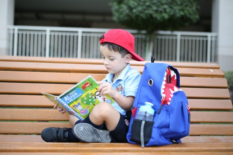 A young boy reads his book on a bench in his school uniform.