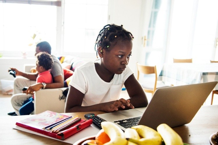 A child uses a laptop at a table while a younger child and a parent sit together on a sofa in the background.