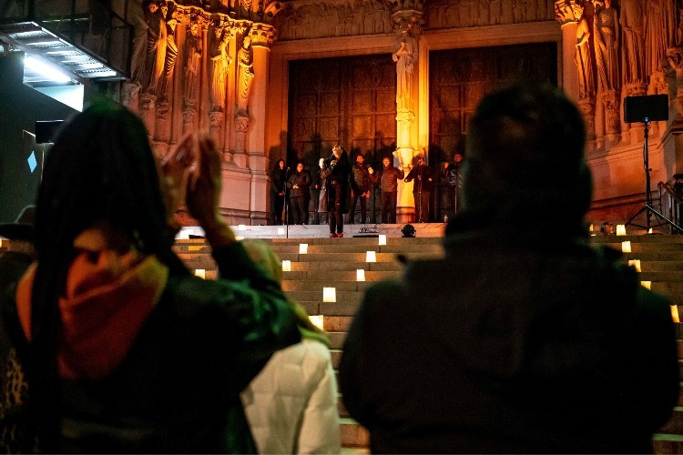 A choir performing at the top of cathedral steps. There are lanterns on the steps. A crowd, backs to the camera, claps for the performance.