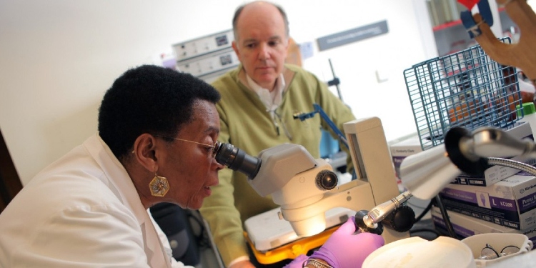 Neurologist looking through a microscope with an elderly man watching from afar.
