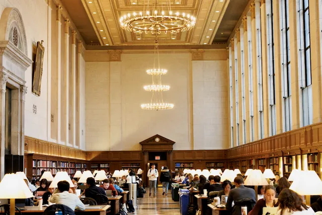 Students fill the tables in the vast main reading room of Columbia University's Butler Library where light pours in from the tall windows and the central chandelier.