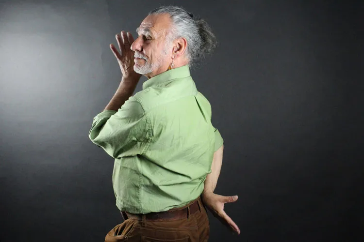 Older man posing for the camera with one hand near his face and the other behind his back. 