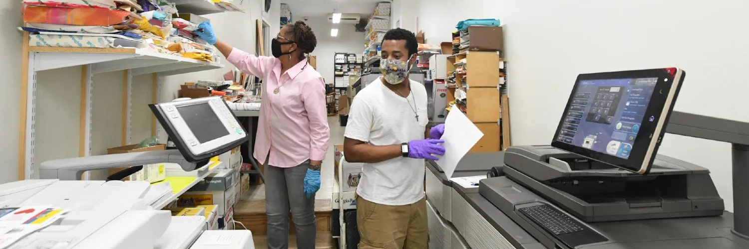 Two people wearing masks stand between two large copiers.
