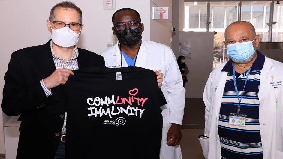 New York City Council member Mark Levine, Dr. Olajide Williams, and Dr. Rafael Lantigua in the lobby of The Forum. Levine holds up a black Community Immunity t-shirt that highlights the word Unity 
