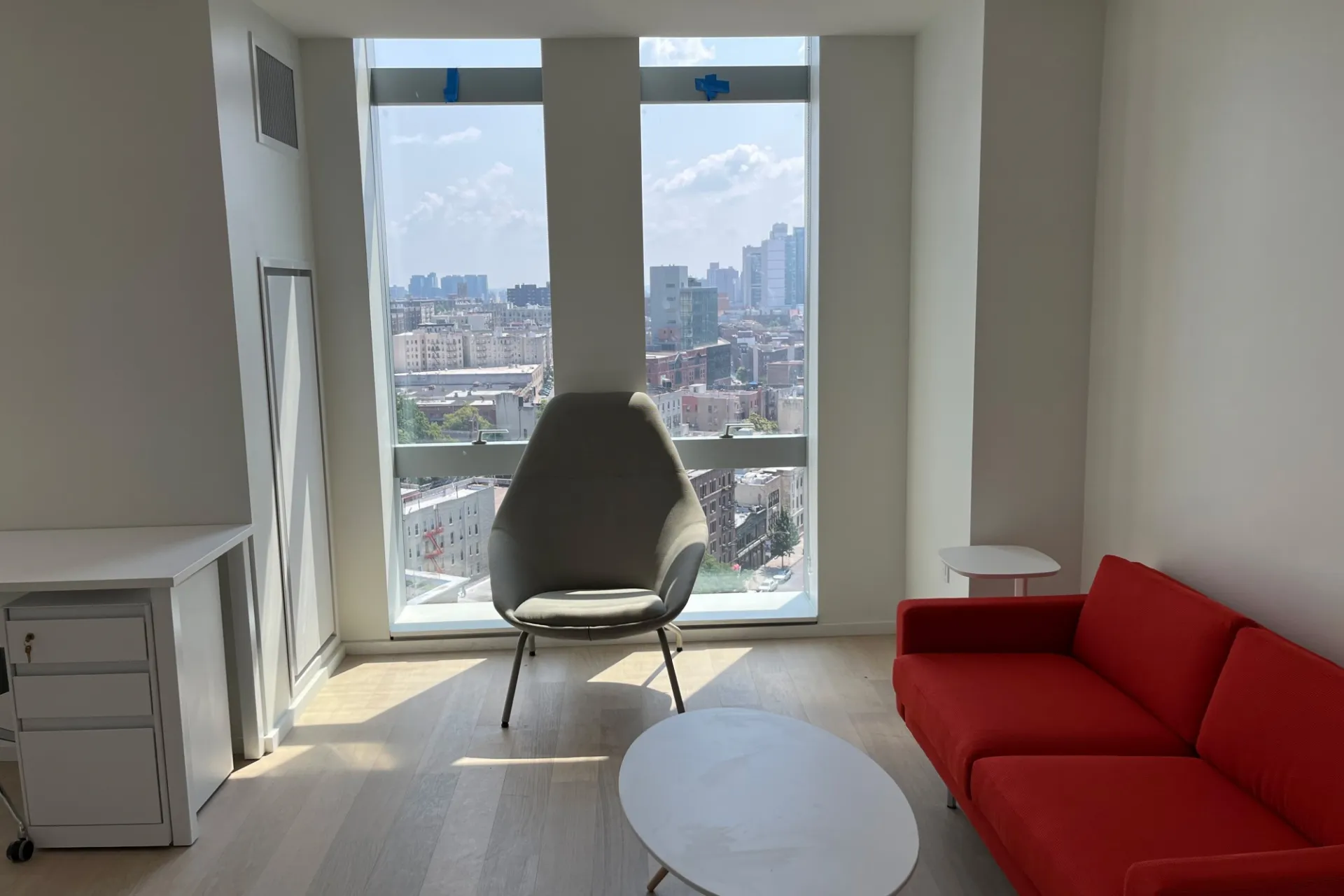 A completed apartment in 600 W. 125th Street that has a view of a desk, couch, coffee table, and chair against a top-to-bottom window that overlooks the city.