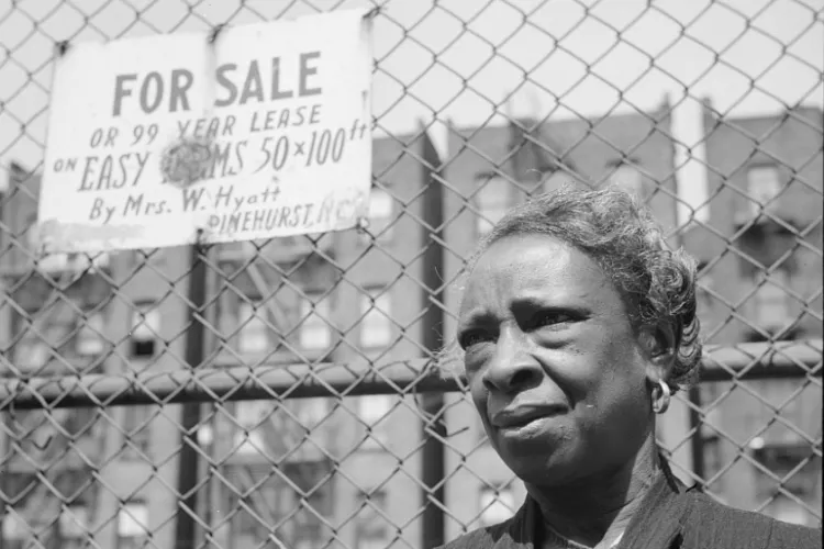 A woman stares out in front of a chain-link fence with a sign reading "FOR SALE"