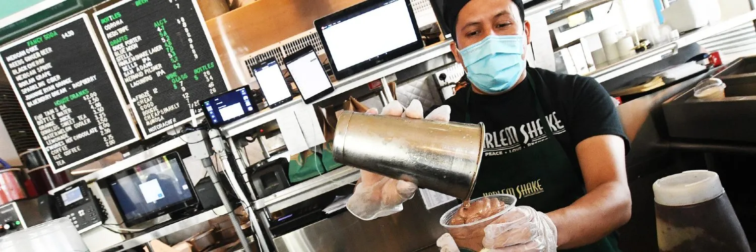 A man wearing a mask and a Harlem Shake shirt and apron pours a chocolate milkshake into a plastic cup that already has vanilla milkshake in it.