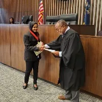 A smiling young woman holding a certificate shakes hands with a judge in a courtroom.
