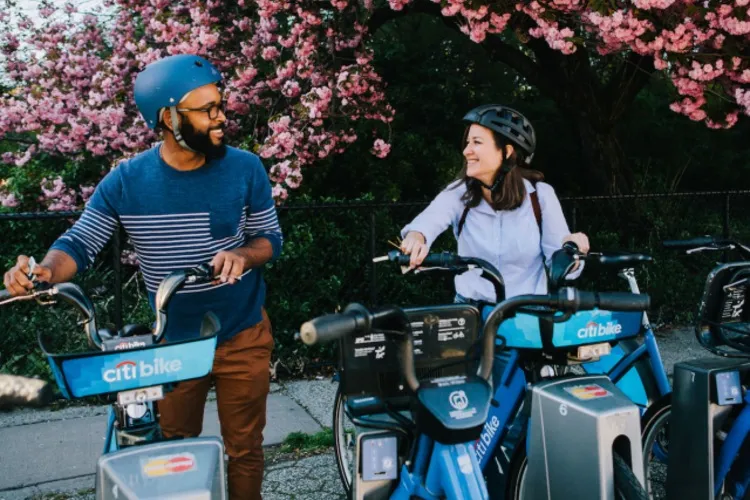 two individuals at a citi bike station, grabbing bikes, smiling, with cherry blossoms in the background.