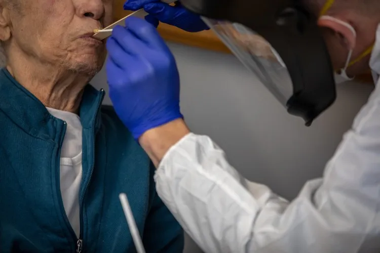 A man has a tongue depressor put in his mouth by a doctor wearing a plastic face shield.