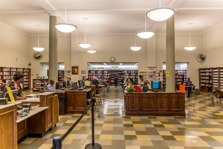 The interior of a library showing the circulation desk, people working at computers, and books along the walls.