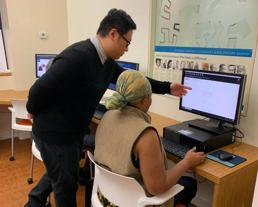A man points to something on the computer screen while a woman sitting at the computer watches.