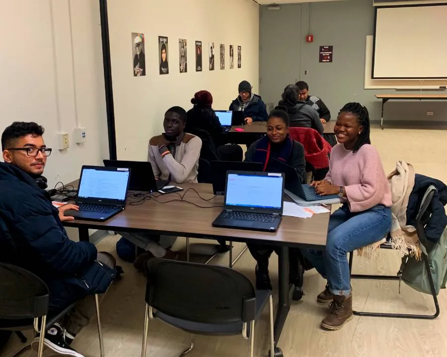 A group of 8 people, mostly young adults, sitting at laptop computers at tables. The 4 in front are looking at the camera and smiling.