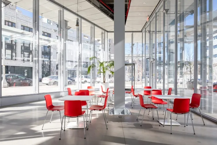 Tables with orange chairs in an airy room with big windows.